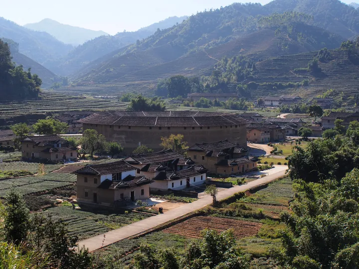 Large circular earthen tulou building surrounded by smaller houses and terraced fields in a mountain valley