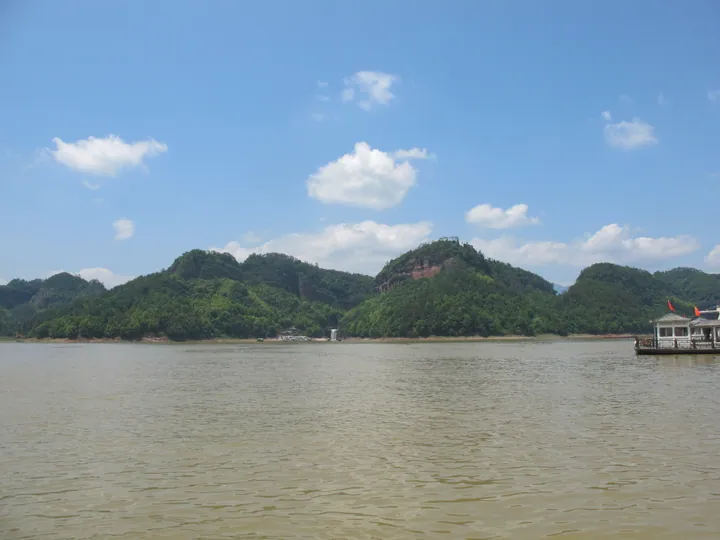 Green forested karst hills beside a lake under a blue sky, with a white tour boat.
