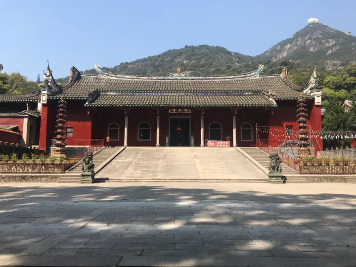 Red temple hall with a tiled roof in front of a forested mountain.