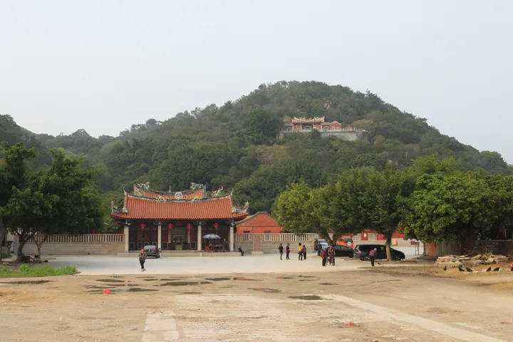 Red-roofed temple building at the base of a green hill with a small pavilion on the hilltop.