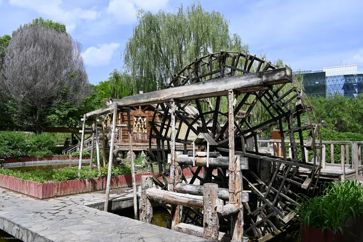 A large wooden waterwheel beside a small pavilion, with trees around