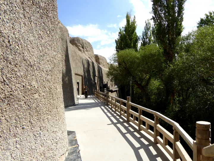 Wooden walkway along a cliff face with carved cave entrances and trees alongside.