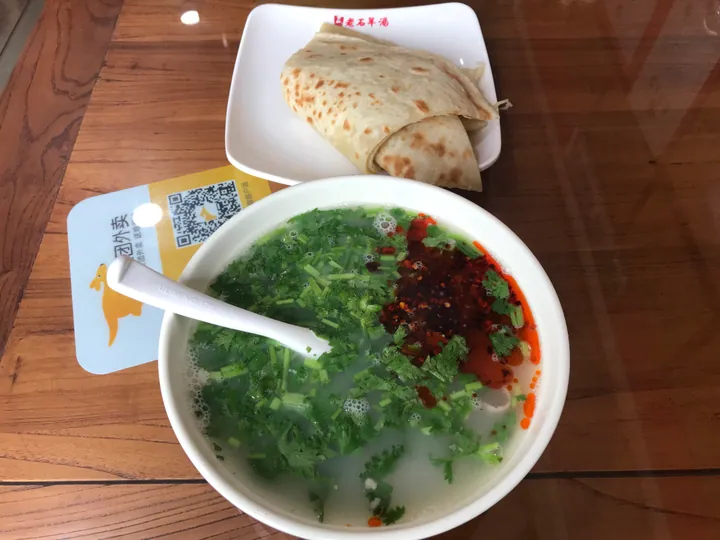 A bowl of mutton offal soup topped with cilantro and chili oil, beside a plate of flatbread