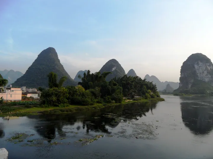 Pointed karst mountains reflected in a calm river with green fields in the foreground