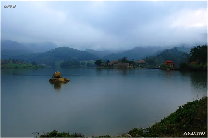 Misty lake surrounded by forested mountains with thatched-roof buildings on the shore