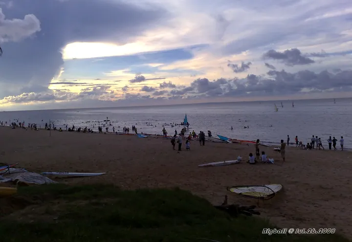 Beach at dusk with people and windsurfing boards on the sand under a colorful sky