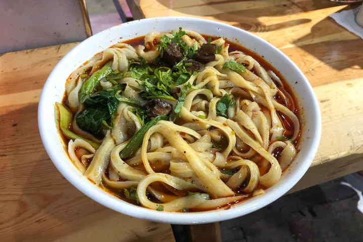 A bowl of wide knife-cut noodles in red broth with meat and herbs