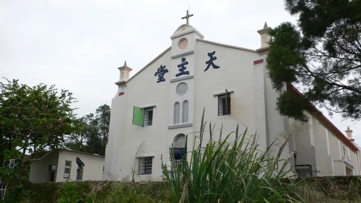 White Catholic chapel with a cross on its facade, surrounded by greenery