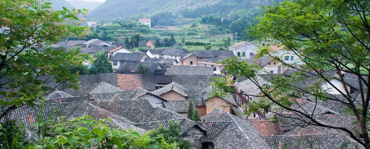 A dense cluster of grey-tiled traditional rooftops in a village nestled among green hills