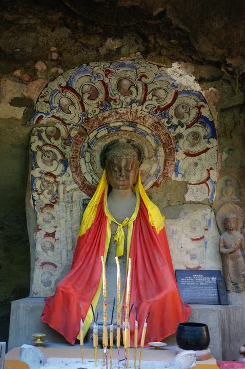 Robed Buddha statue with a painted halo carved into a cave wall, with incense sticks in front