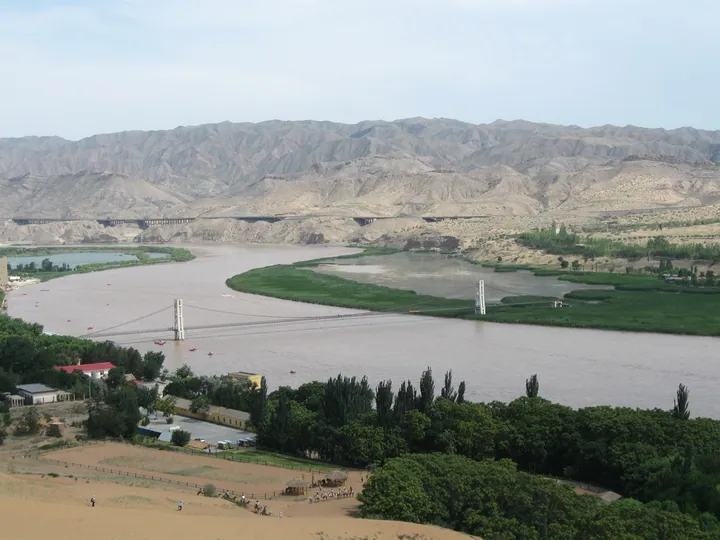 A river bend with a suspension bridge, green wetland, and desert dunes