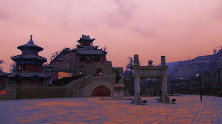 Palace complex on a hillside at dusk with a memorial arch and statue in the foreground plaza