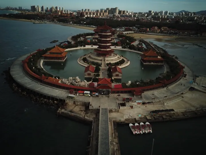 Aerial view of a red pavilion complex surrounded by a circular moat, with the city skyline behind