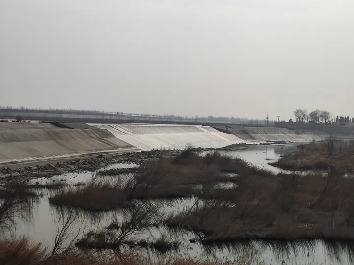 Long sloped stone dam spillway beside a shallow river with dry reeds in the foreground.