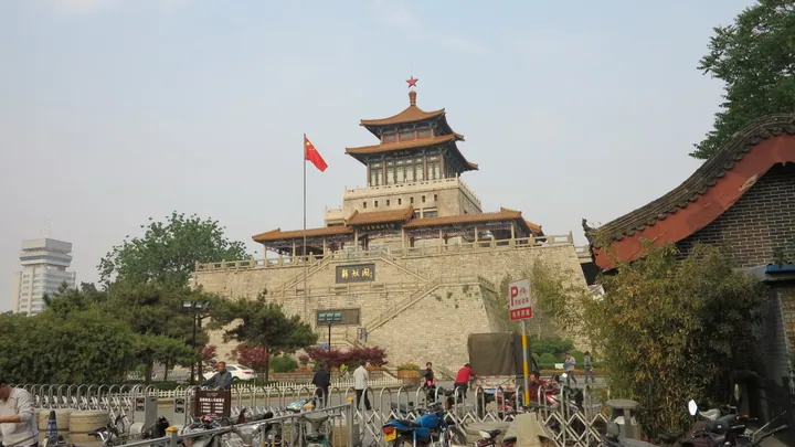 Multi-tiered Chinese pavilion on a brick city-wall platform with a red flag in front.