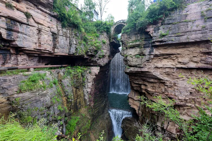 A narrow rock canyon with a waterfall and a stone arch bridge overhead