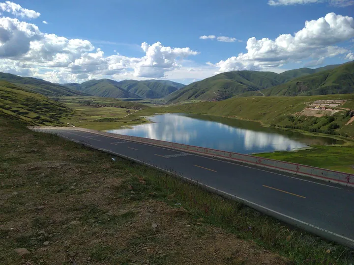 A calm blue lake reflecting clouds, framed by green mountains and a road