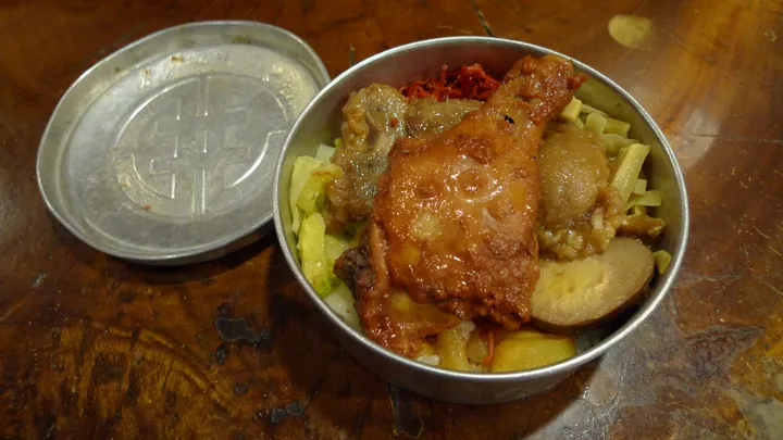 An open lunch box with a chicken leg, braised items, and pickled vegetables over rice.