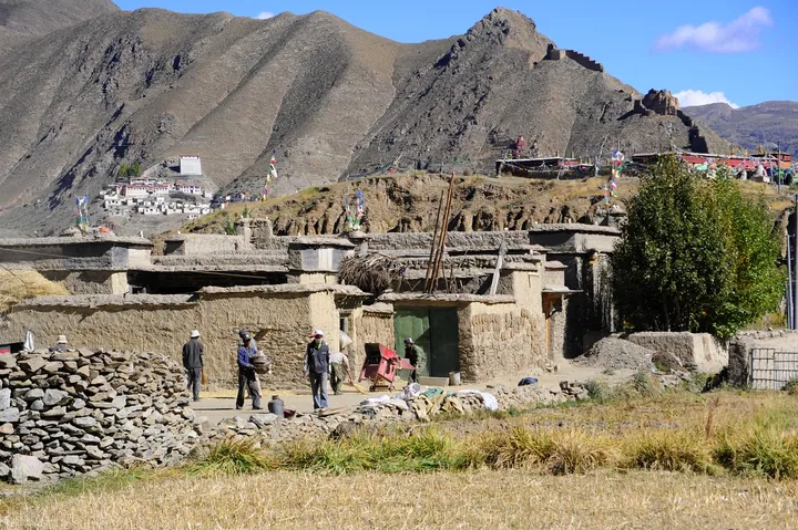 Mud-brick village buildings in a valley with mountains and a flag-covered hillside in the background.