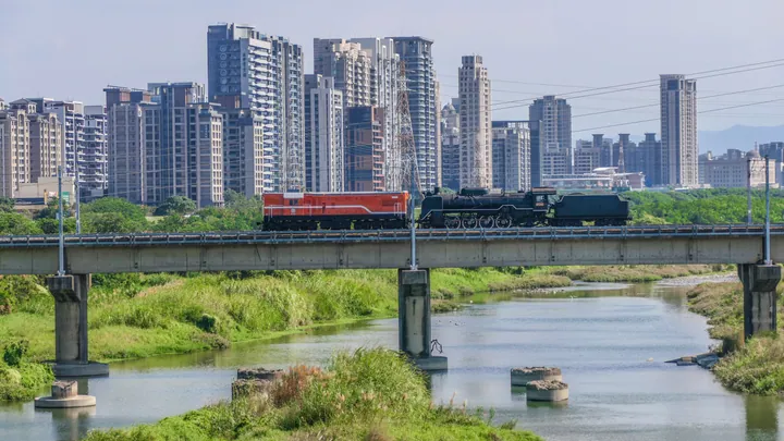 A steam locomotive crossing a bridge with the Zhubei skyline behind it