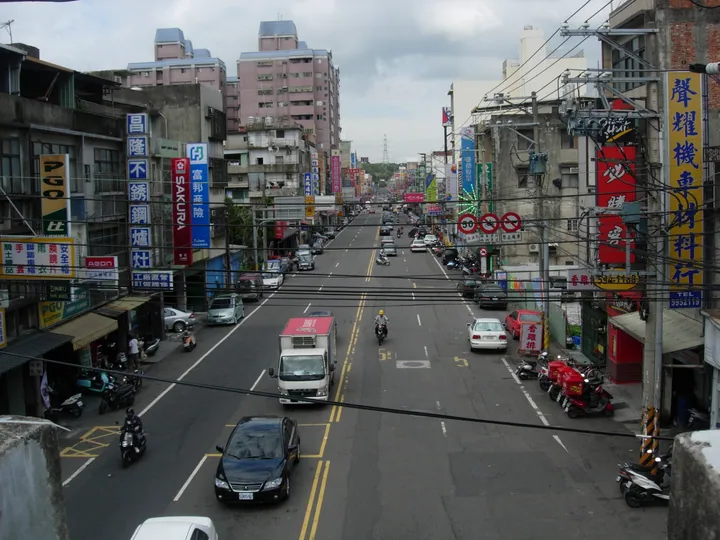 A busy street lined with shop signs, cars and scooters, with apartment buildings in the background
