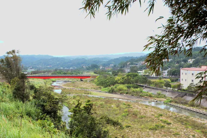 A red bridge crossing a stream, with hills and houses in the background