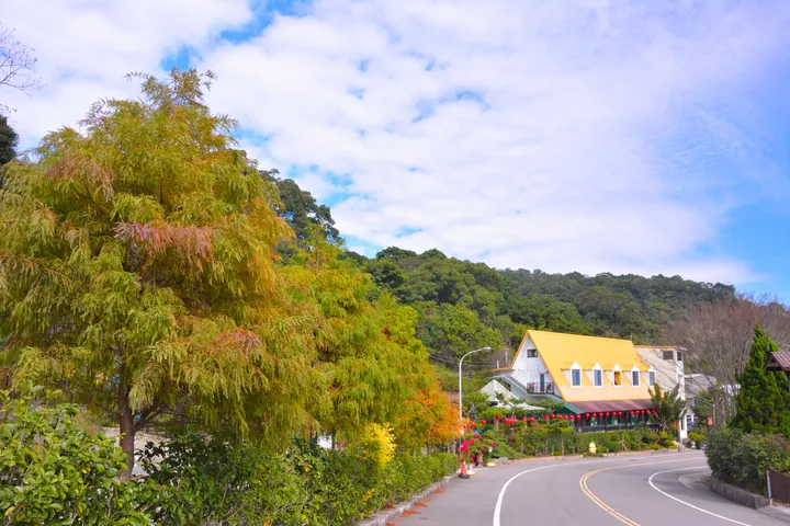 A curving road past a chalet-style yellow-roofed building surrounded by autumn-coloured trees under a blue sky