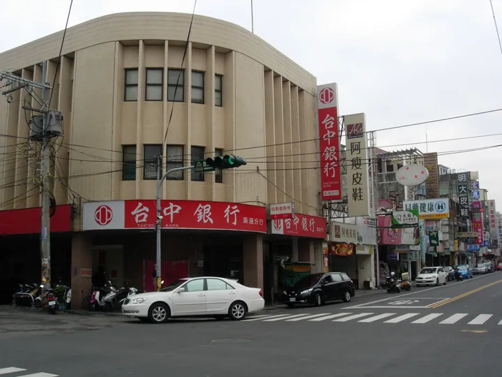 A bank building on a street corner with shops, cars and a traffic light