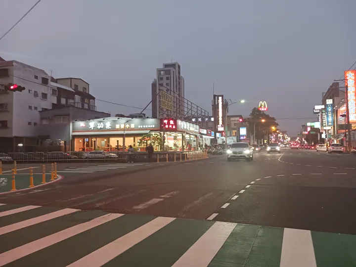Dusk street intersection with illuminated restaurant signs and passing cars in Taiping District, Taichung