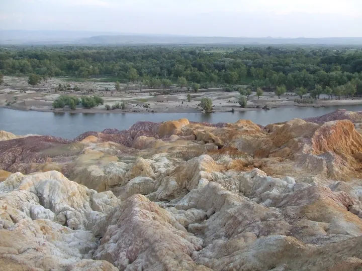 Multicoloured eroded rock formations along a river beneath a forested far bank