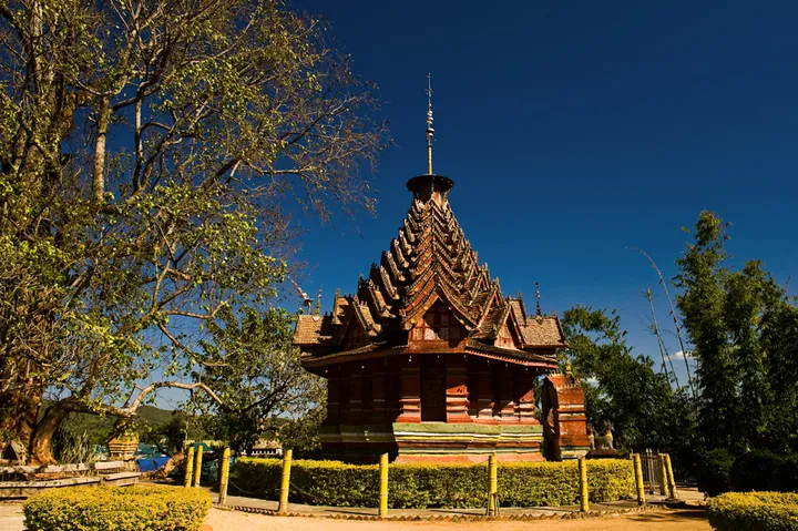An ornate multi-tiered wooden octagonal pavilion with a spire, set among trees under a blue sky