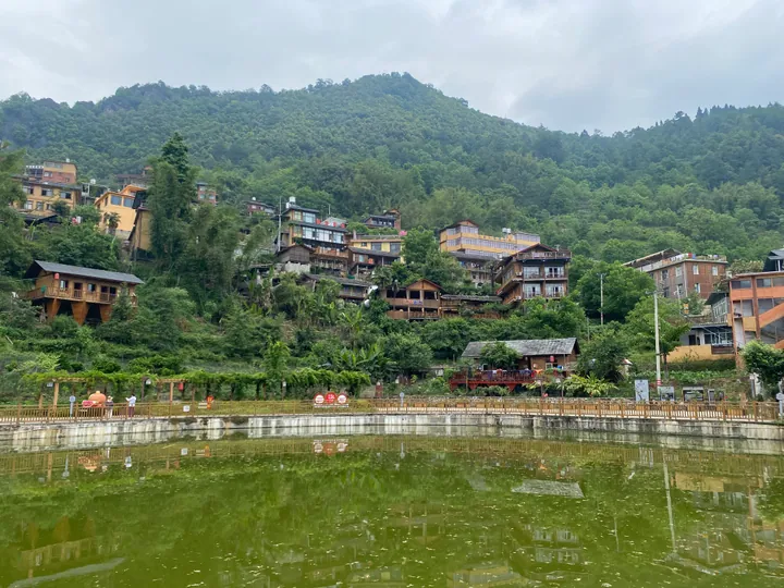 Hillside village with wooden houses overlooking a green pond, surrounded by forested mountains