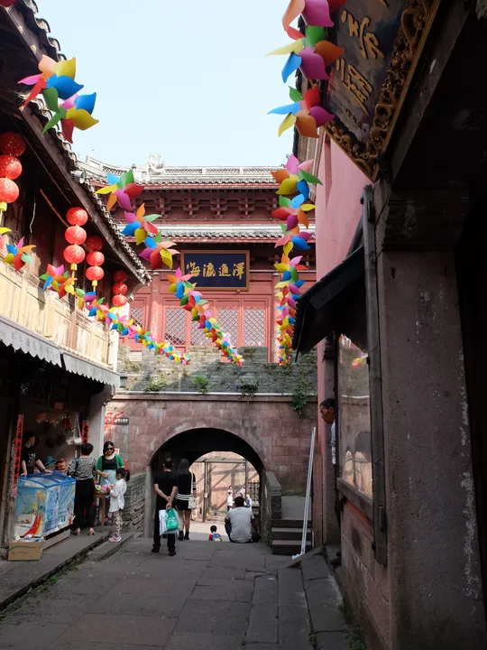 Narrow stone alley strung with colourful paper decorations and lanterns leading to an arched gate in the old fishing town