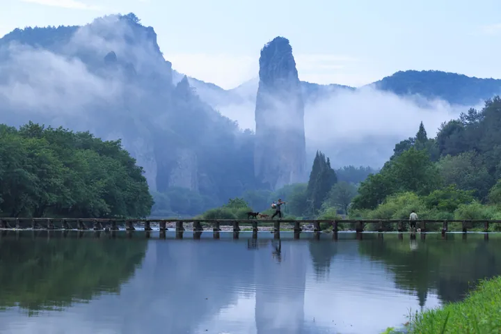 A tall rock pillar reflected in a calm river with a wooden bridge in the foreground and misty mountains behind.