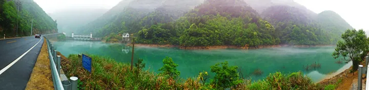 Panoramic photo of a turquoise reservoir surrounded by misty green mountains.