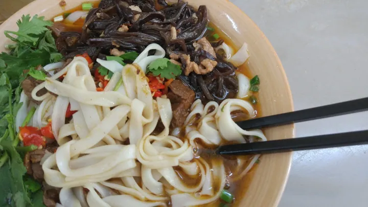 A bowl of wide rice noodles in broth with beef, black fungus, chili and cilantro