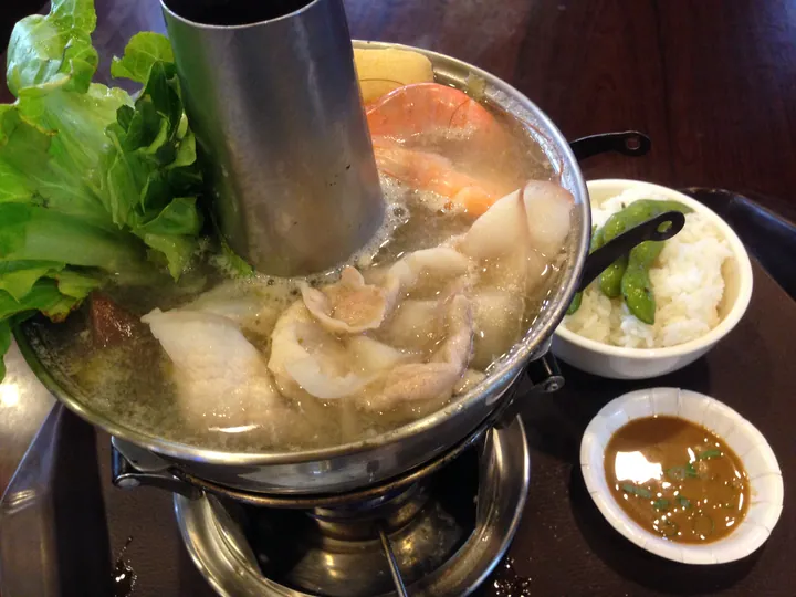 A steaming hot pot of sauerkraut, sliced pork and seafood with a side of rice and dipping sauce