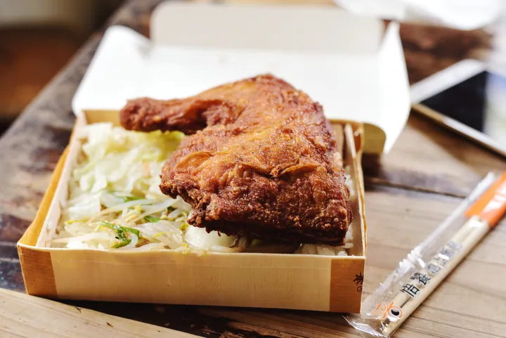 A wooden lunch box holding fried chicken over rice with vegetables and chopsticks.