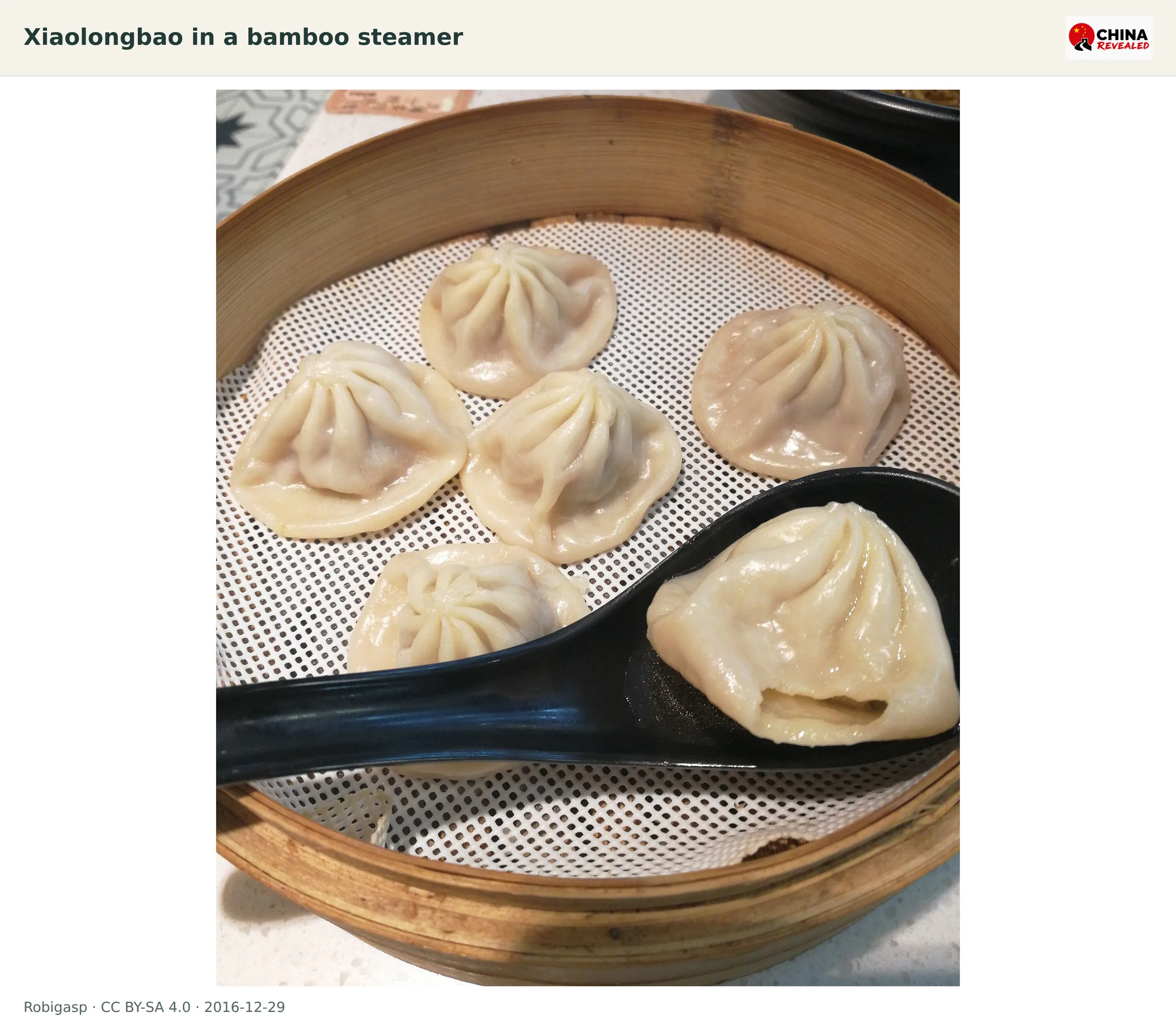 Several Shanghai-style soup dumplings in a bamboo steamer with one dumpling opened on a spoon